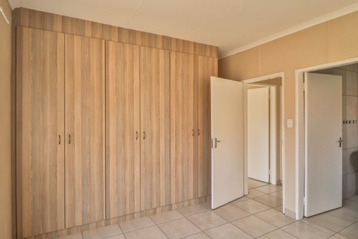 A clean, empty bedroom featuring light wood-finish built-in cupboards, neutral tiled flooring, and access to adjacent rooms.
