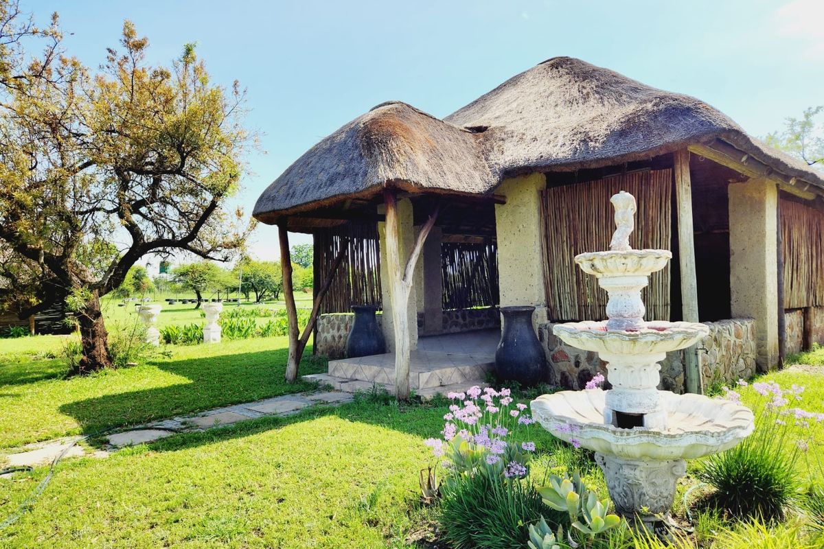 A charming outdoor thatched-roof gazebo featuring stone masonry, timber supports, and a decorative fountain set within a well-maintained garden landscape.