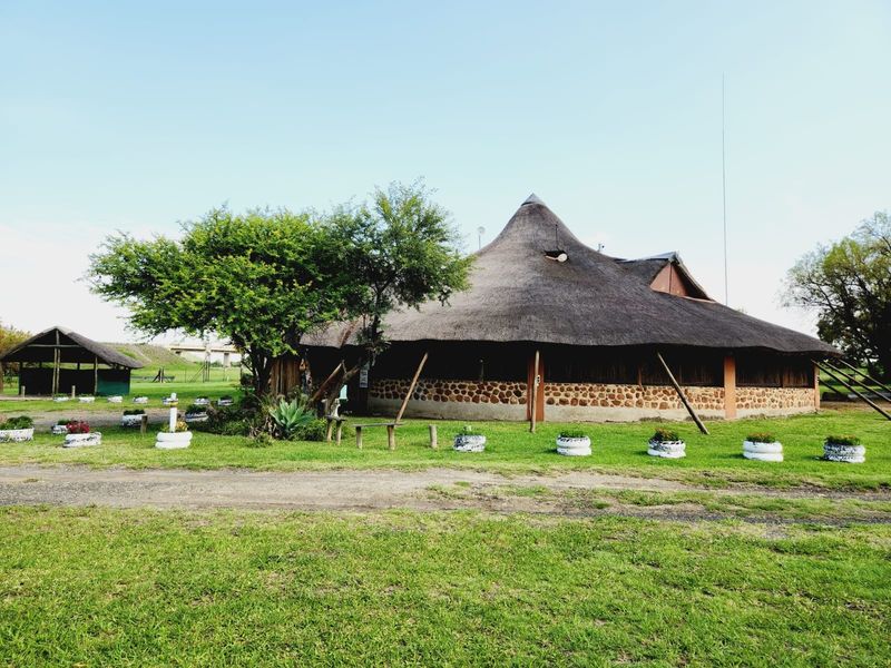 A rustic farm property featuring a large thatched-roof building with stone masonry walls and a separate open-sided outbuilding set in a grassy landscape.