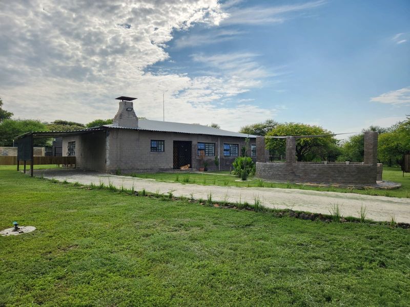 Rear view of a farm property featuring a corrugated metal roof, brick chimney, covered patio area, and a stone masonry structure set in a garden.