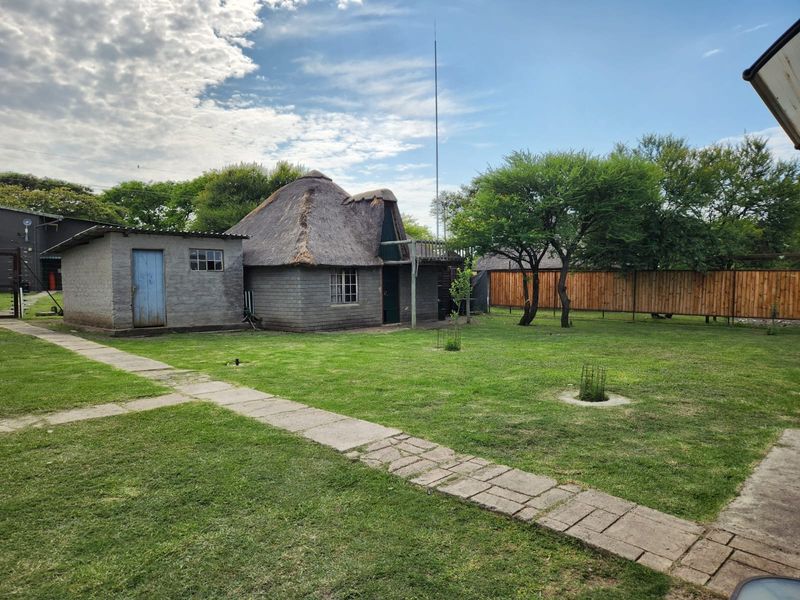 A rear view of a farm property featuring a traditional thatch-roofed structure, a secondary outbuilding, and a spacious garden with a paved walkway.