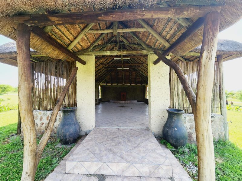 An open-air thatched lapa or patio area featuring rustic timber supports, stone masonry, and tiled flooring, ideal for outdoor entertaining on the farm.