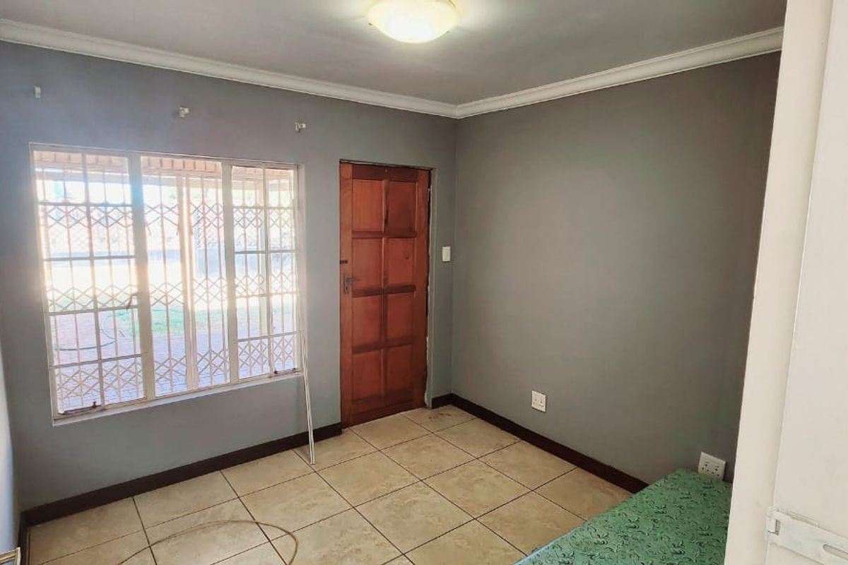 A functional bedroom featuring tiled flooring, a large window with security bars, and a wooden door, providing a neutral base for personalization.