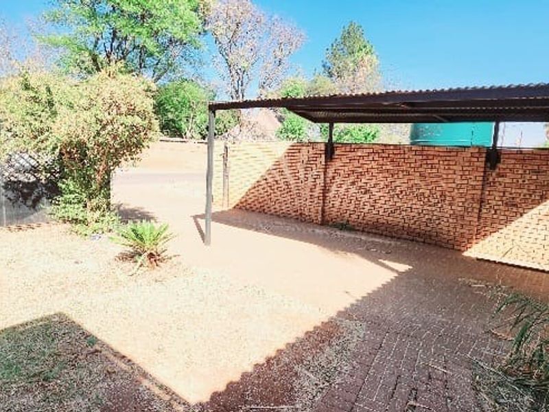 Outdoor area featuring a paved driveway, a covered carport structure, and a brick boundary wall with established garden surroundings.