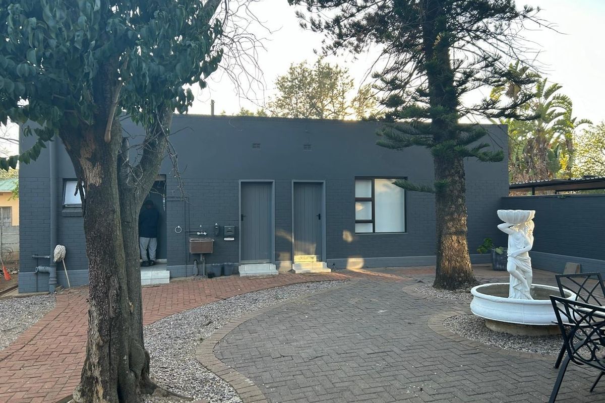 A rear view of the property featuring a dark-painted brick exterior, paved courtyard area, and established trees.