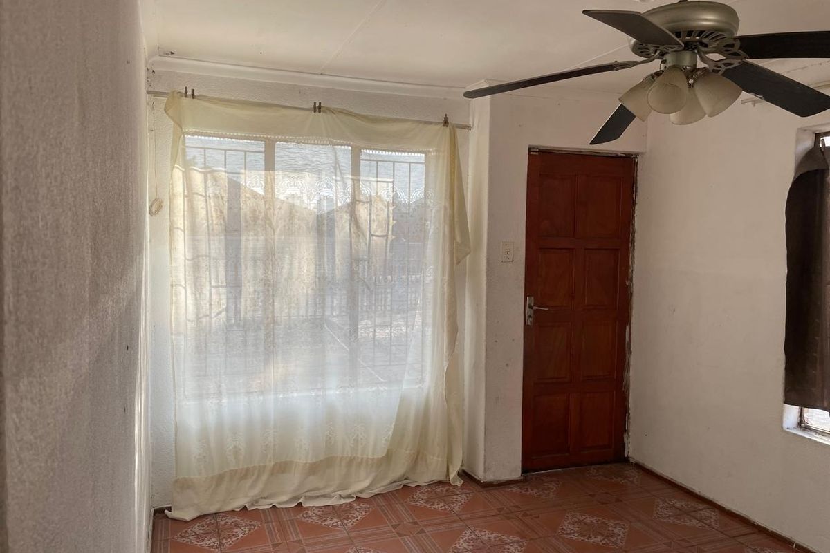 A living room featuring tiled flooring, a ceiling fan, and a wooden door, with large windows fitted with security bars.