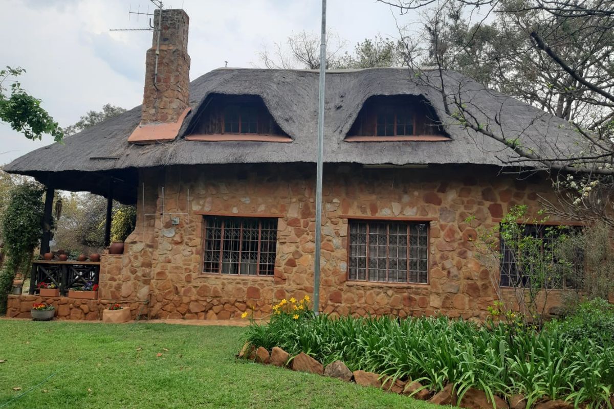 A charming stone-clad farmhouse featuring a traditional thatched roof, dormer windows, and a prominent stone chimney, set within a well-maintained garden.