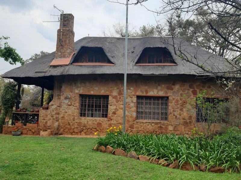 A charming stone-clad farmhouse featuring a traditional thatched roof, dormer windows, and a prominent stone chimney, set within a well-maintained garden.