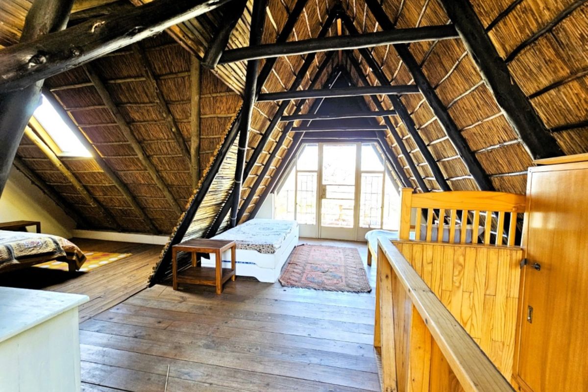 A spacious loft bedroom featuring exposed timber beams, a traditional thatched roof, wooden flooring, and access to a balcony through glass doors.