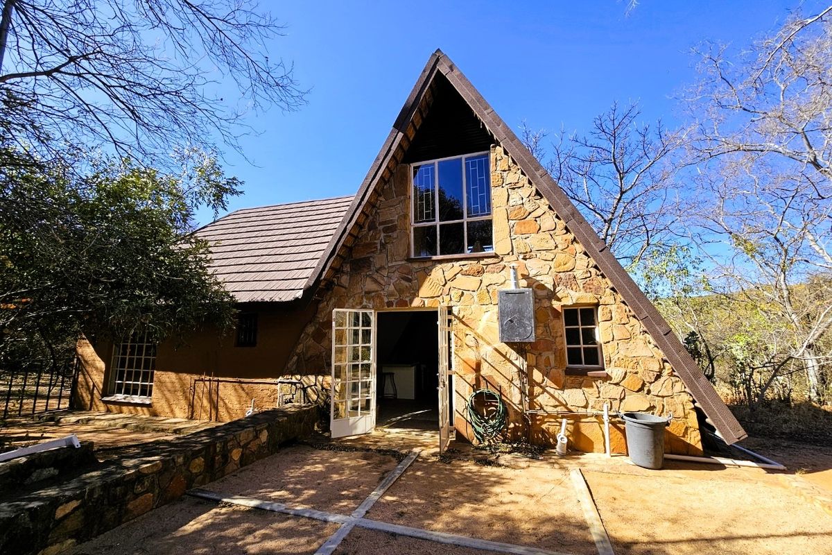 A-frame style house featuring natural stone cladding, tiled roof, and a paved patio area set within a natural, wooded environment.