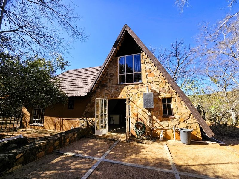 A-frame style house featuring natural stone cladding, tiled roof, and a paved patio area set within a natural, wooded environment.