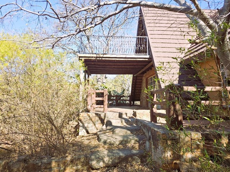 A rustic A-frame house exterior featuring a covered patio, stone retaining walls, and an elevated balcony with decorative metal railings.