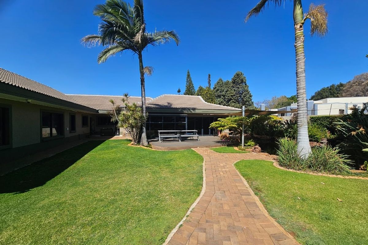 A well-maintained rear garden featuring a paved walkway, lush lawn, mature palm trees, and a wooden deck area attached to the house.