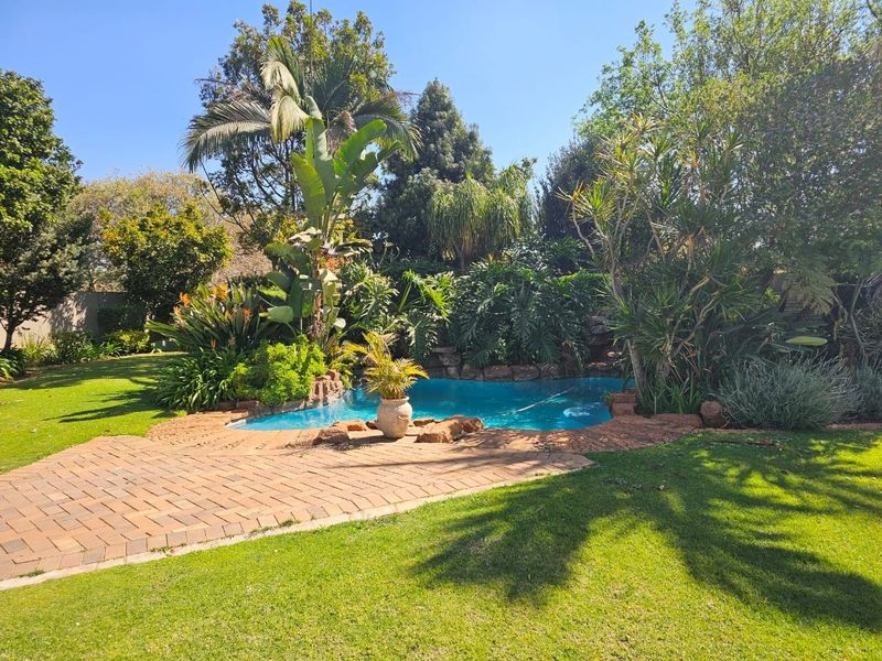 A well-maintained swimming pool area featuring a brick-paved surround, lush established garden landscaping, and mature trees in a private backyard setting.