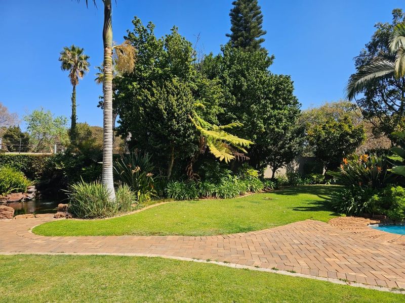 A well-maintained garden area featuring a paved walkway, lush green lawn, mature trees, and a glimpse of a swimming pool.