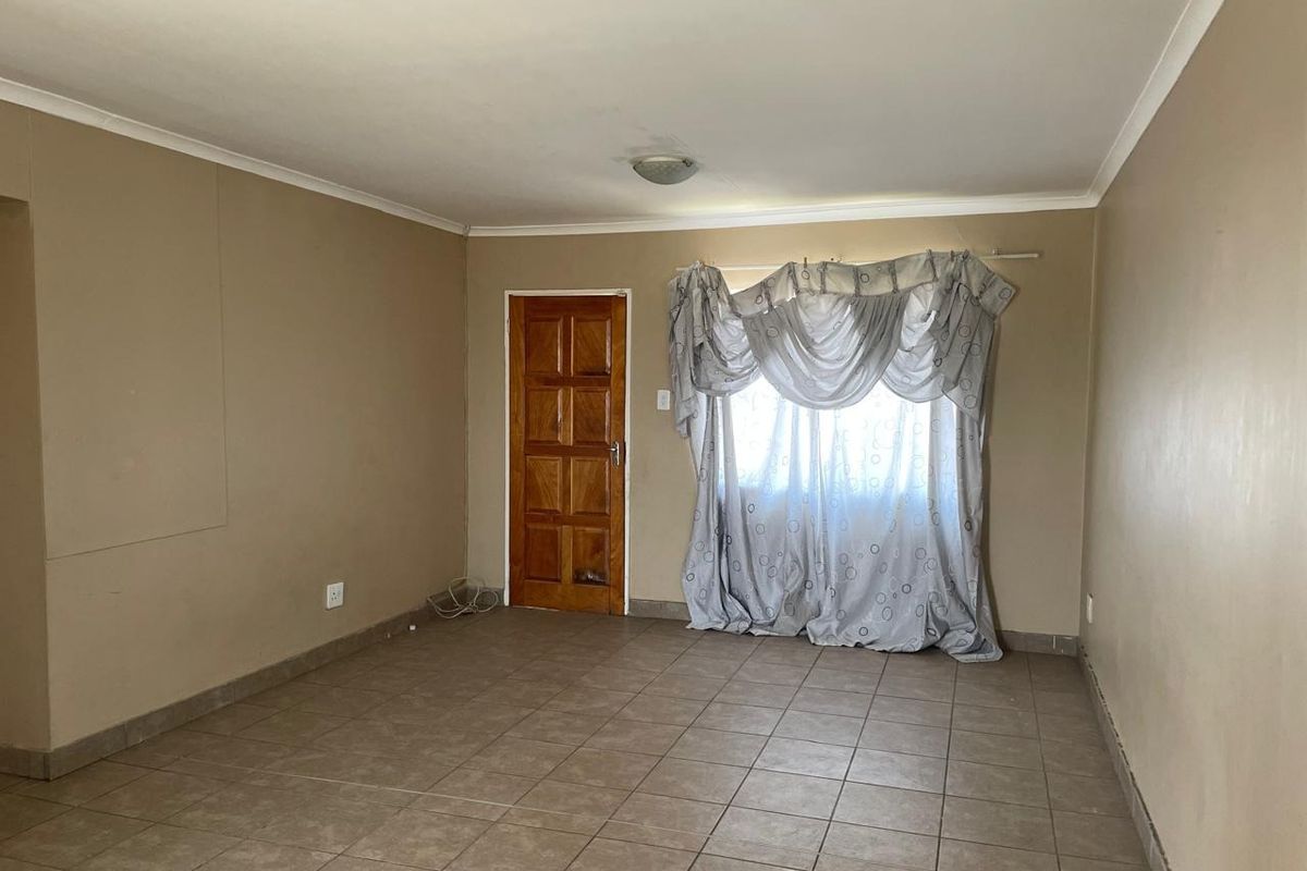 A spacious living room featuring neutral wall tones, tiled flooring, and a wooden door, offering a blank canvas for personalization.