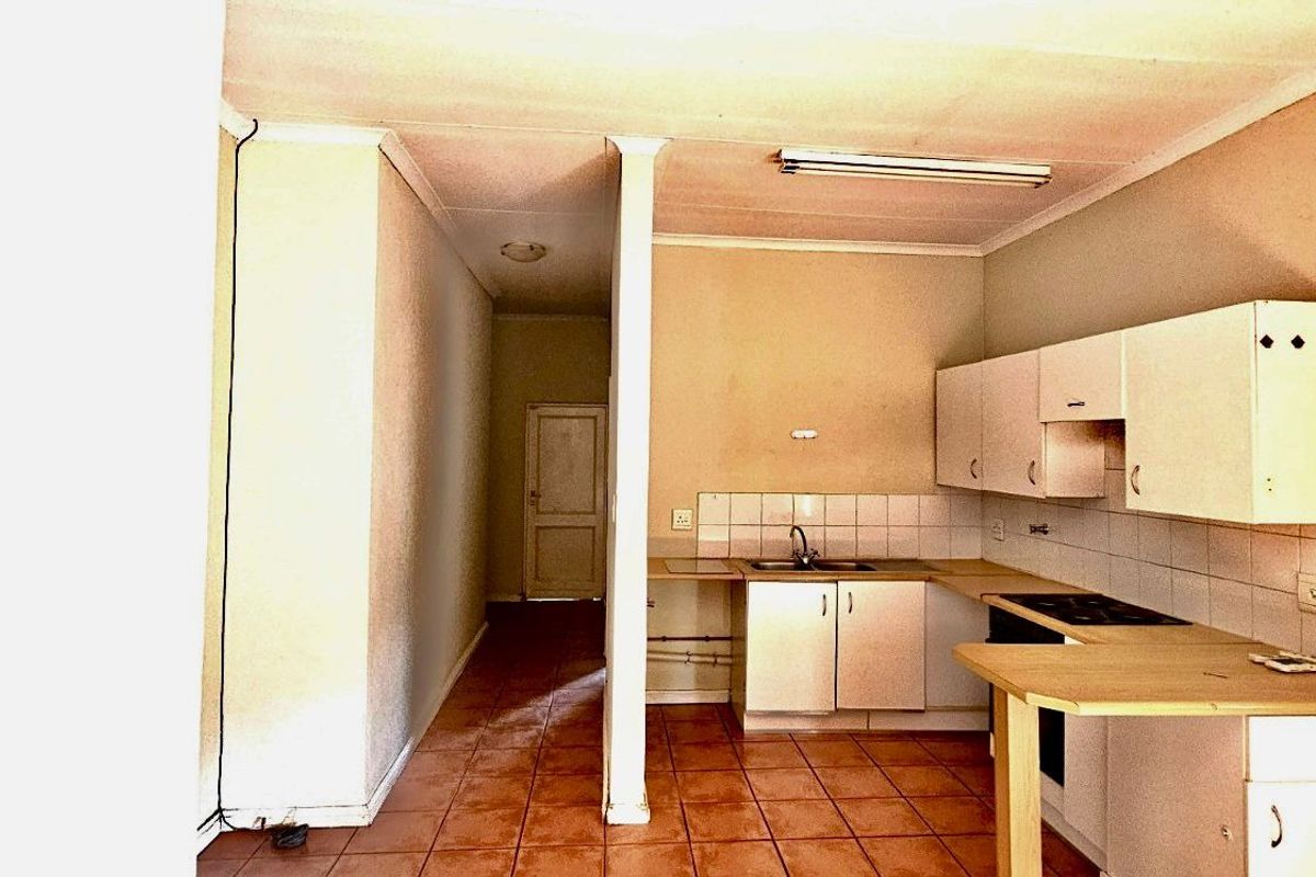 A dated kitchen area featuring basic cabinetry, tiled splashbacks, and a breakfast counter, leading into a hallway with tiled flooring.