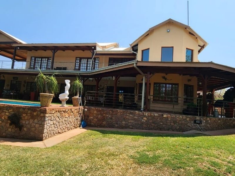 Rear view of a multi-level farm residence featuring a stone-clad base, expansive covered patio, and an integrated swimming pool set within a landscaped garden.
