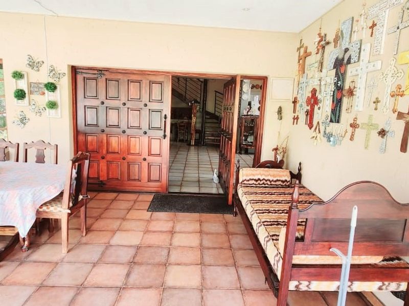 A covered patio area featuring terracotta floor tiles and a large wooden entrance door leading into the main residence.
