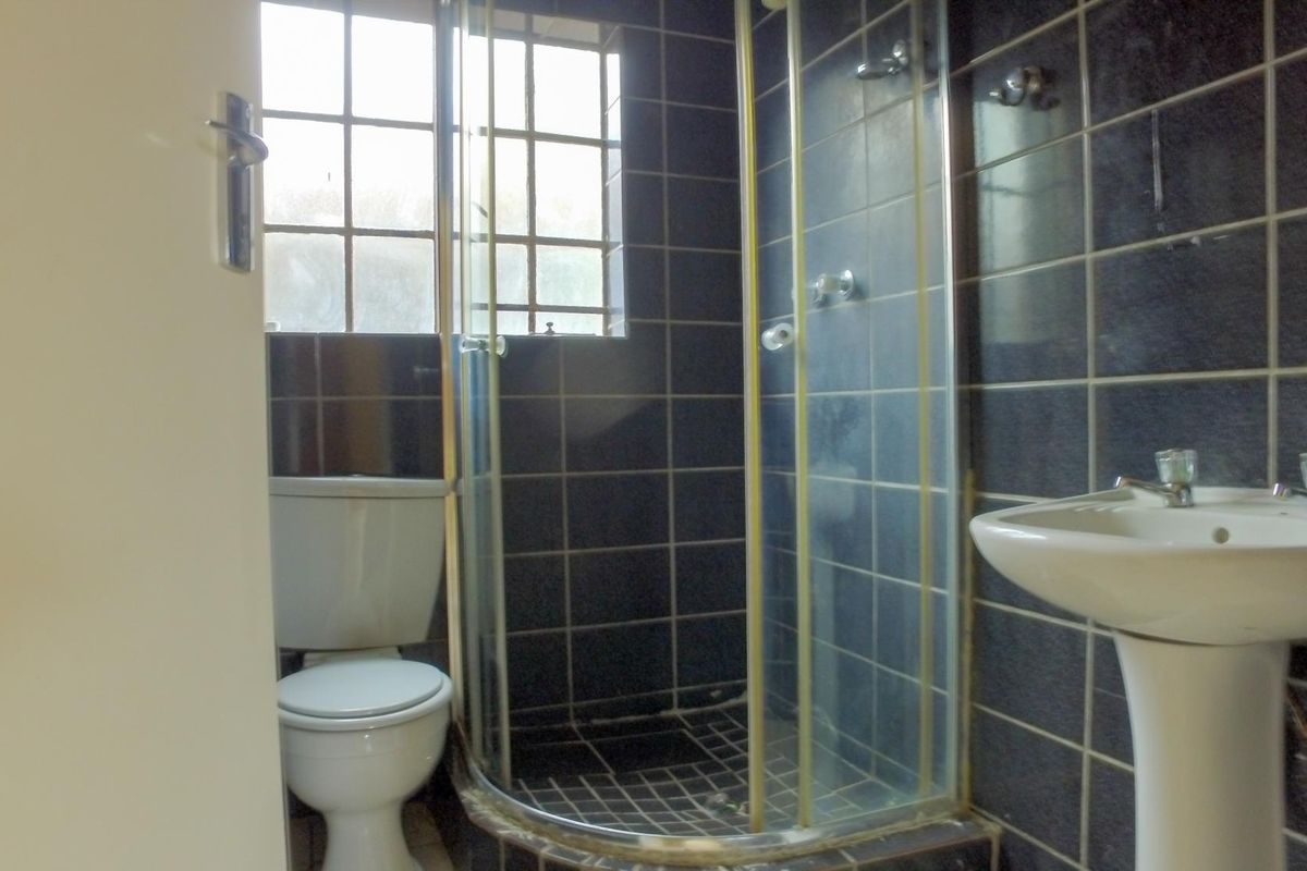 A compact bathroom featuring a corner shower enclosure, pedestal basin, and toilet, finished with dark wall tiling and a window for natural light.
