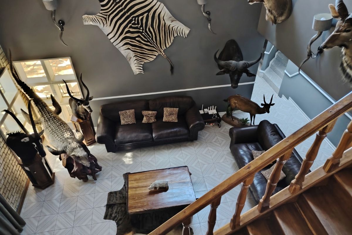 High-ceiling living room featuring tiled flooring, wall-mounted lighting fixtures, and a wooden staircase with a balustrade overlooking the main seating area.