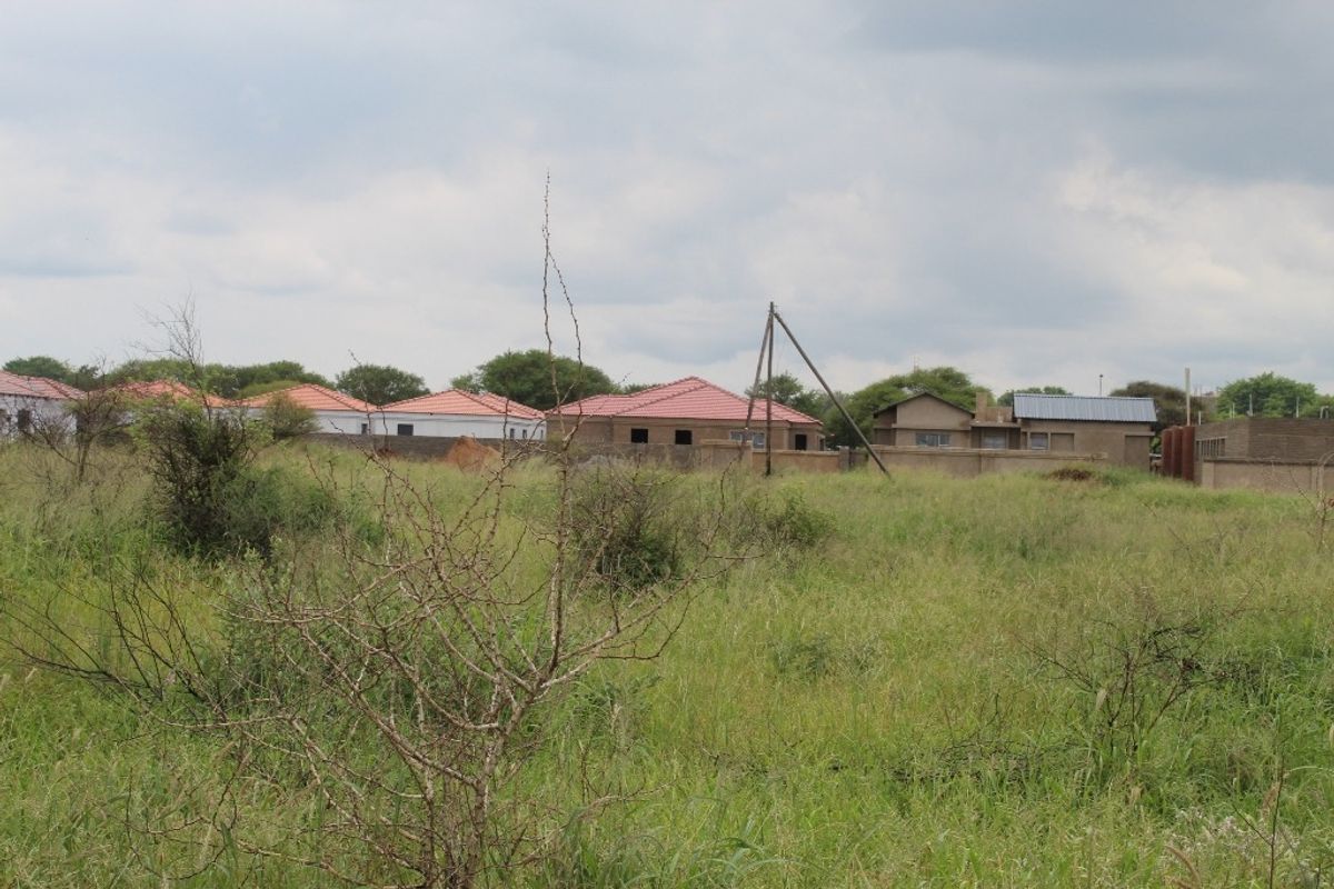 A view of a vacant residential plot featuring natural grass cover with surrounding residential properties in the background.