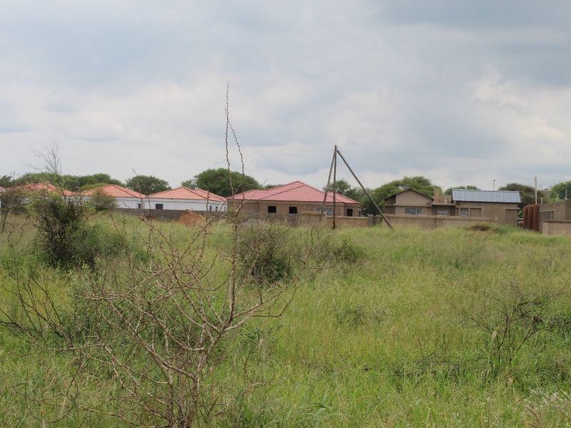 A view of a vacant residential plot featuring natural grass cover with surrounding residential properties in the background.