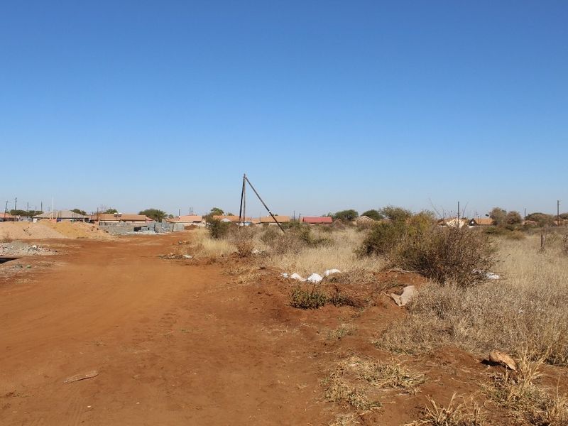 A view of a vacant residential plot featuring natural vegetation, dirt access path, and surrounding neighborhood infrastructure.