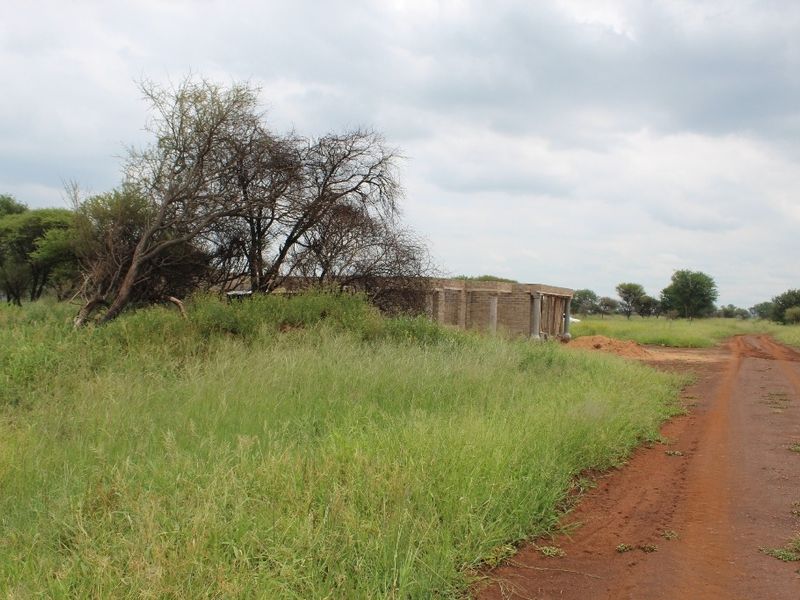 A view of a vacant residential plot featuring an unfinished building structure surrounded by natural vegetation and a dirt access road.