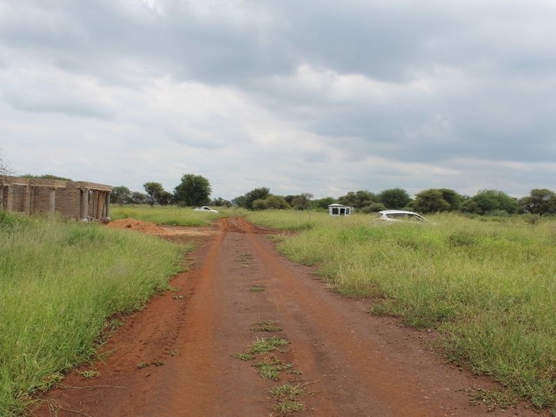 A view of a vacant residential plot featuring an unpaved access road and an unfinished brick building structure set within a grassy landscape.