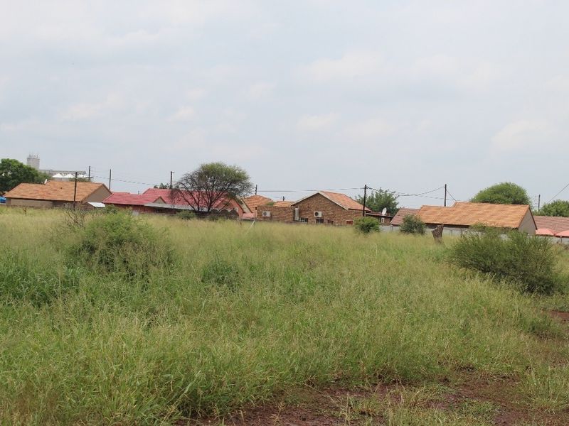 A view of a vacant residential plot featuring natural grass cover with surrounding established residential properties in the background.