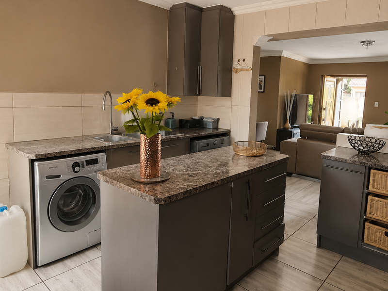 Open-plan kitchen area featuring modern grey cabinetry, laminate countertops, tiled flooring, and integrated laundry appliance space, flowing into the adjacent living room.