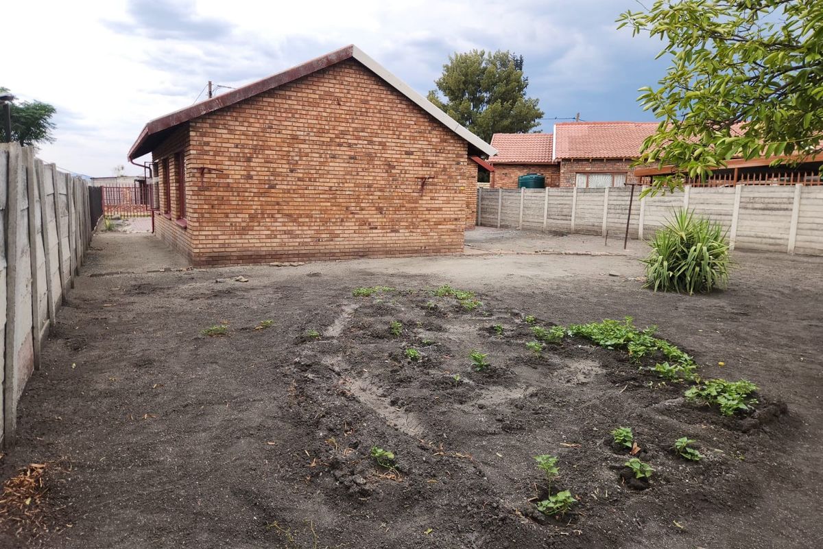 A view of the rear garden area featuring a brick exterior wall, perimeter concrete walling, and an unpaved yard with a small vegetable patch.
