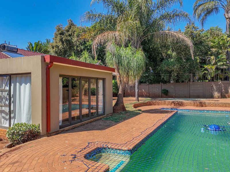 A rear garden area featuring a swimming pool with safety net, brick paved patio, and a detached structure with large glass sliding doors.