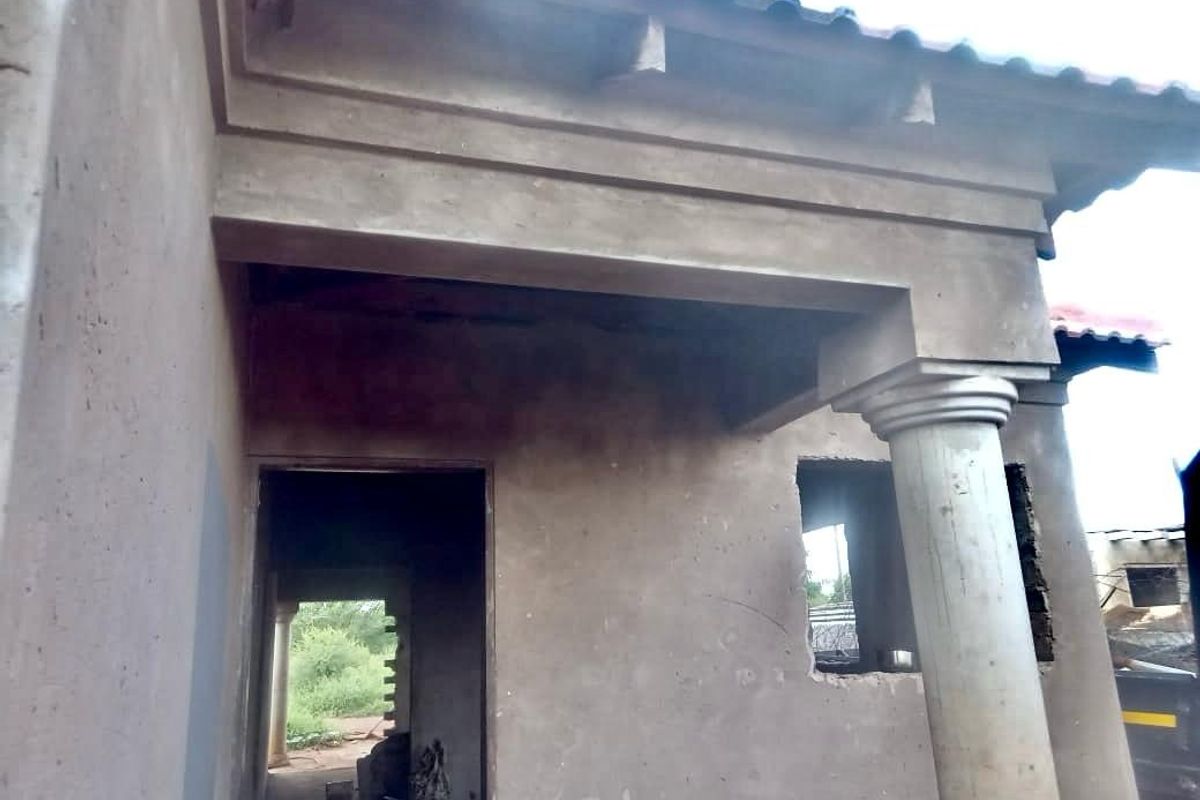 Exterior view of a house under construction, featuring a covered entrance porch with a structural column and unfinished cement plaster walls.
