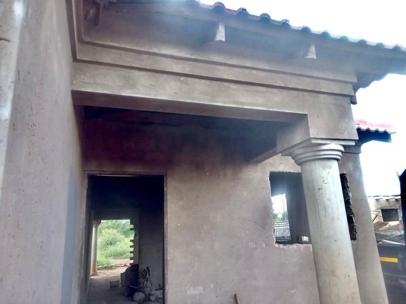 Exterior view of a house under construction, featuring a covered entrance porch with a structural column and unfinished cement plaster walls.
