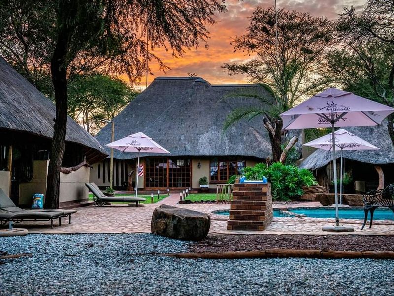 A scenic outdoor pool area featuring traditional thatched-roof structures, a brick braai, and a paved patio set against a beautiful sunset backdrop.