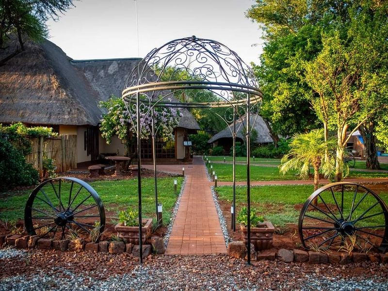 A well-maintained farm garden featuring a brick pathway, a decorative metal gazebo, and traditional thatched-roof structures in a lush, landscaped setting.