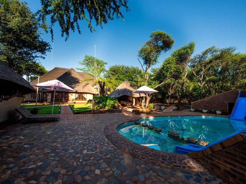 A well-maintained outdoor pool area featuring a water slide, stone paving, and traditional thatched-roof structures set within a lush garden environment.