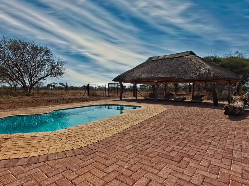Outdoor pool area featuring a swimming pool, brick paved surround, and a traditional thatched lapa structure set within a farm landscape.