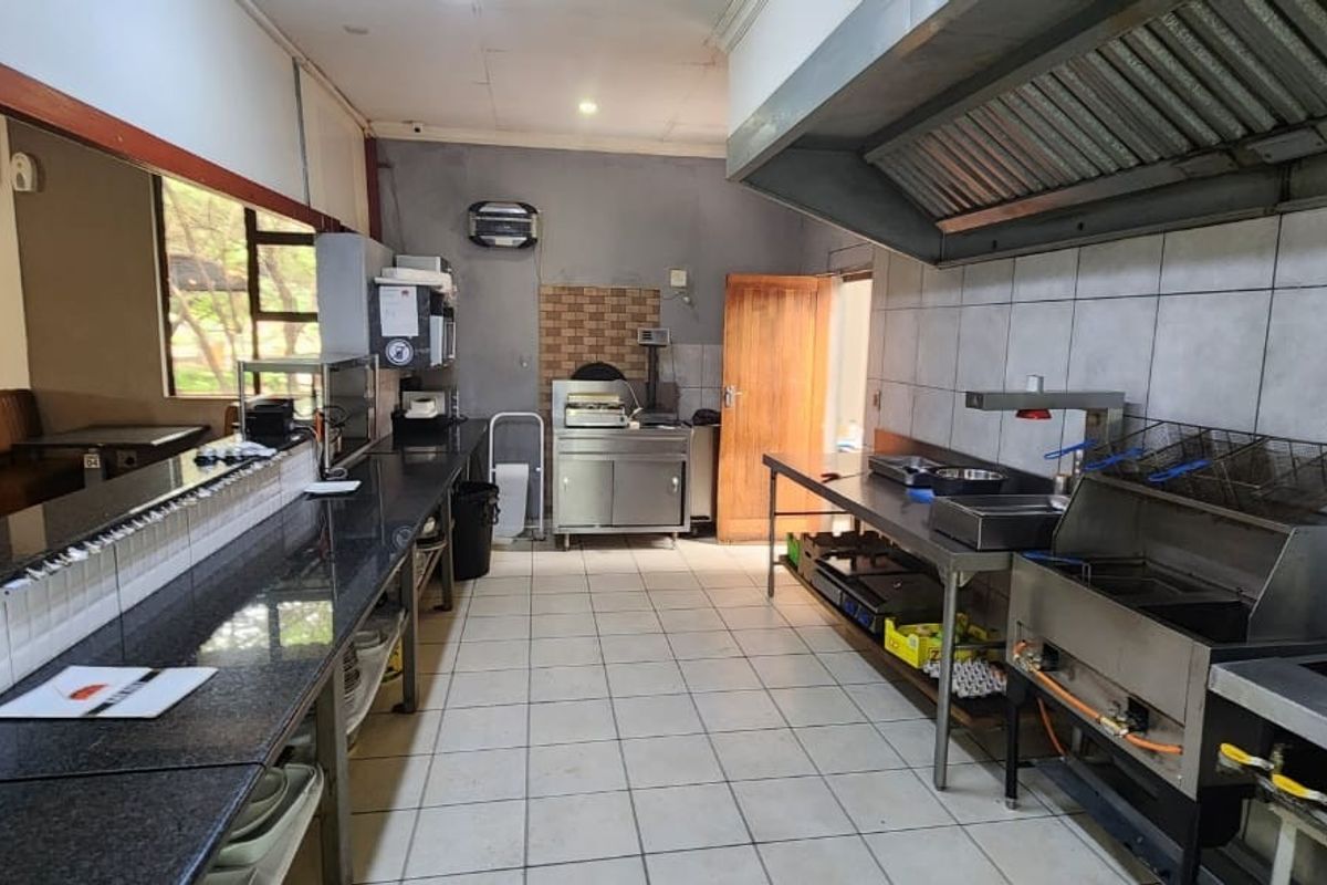 Commercial kitchen area featuring stainless steel prep surfaces, a large extractor hood, tiled walls, and a service window connecting to the dining space.