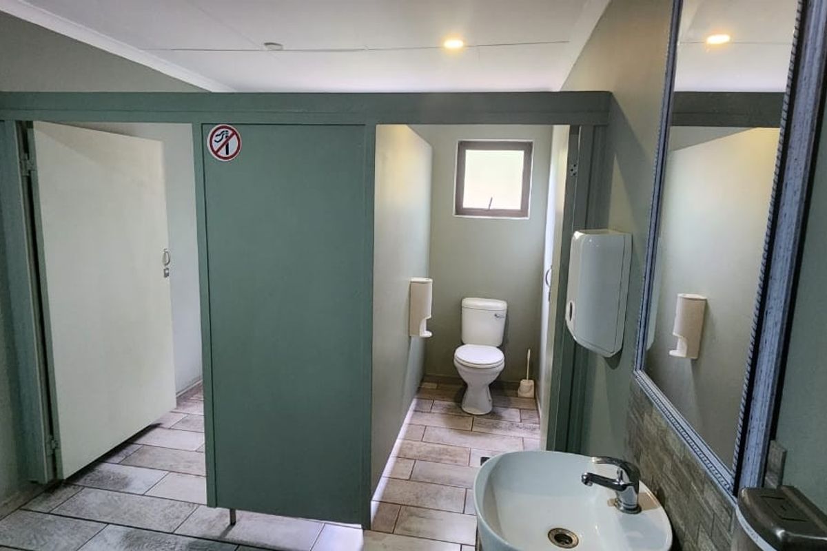 A functional communal bathroom featuring tiled flooring, a private toilet cubicle, and a wall-mounted washbasin with a mirror.