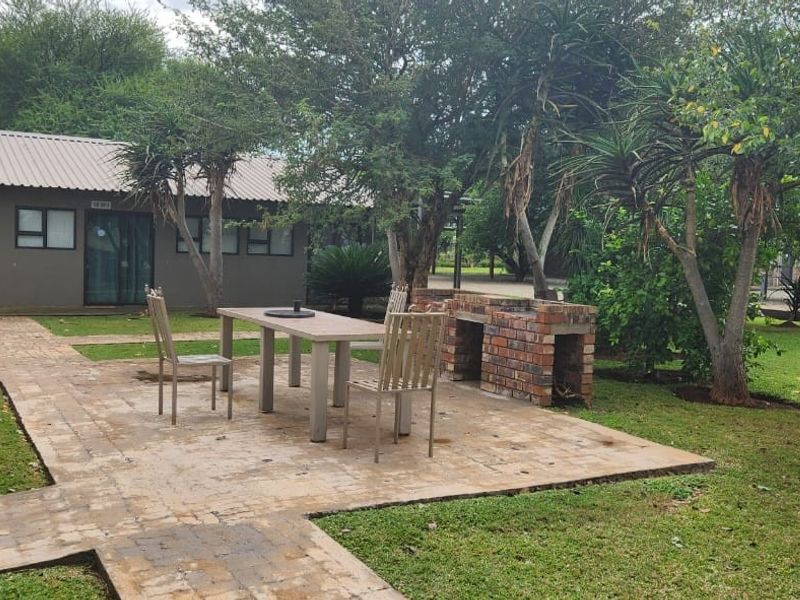 A well-maintained rear garden area featuring a paved patio with a built-in brick braai, surrounded by established trees and lawn.