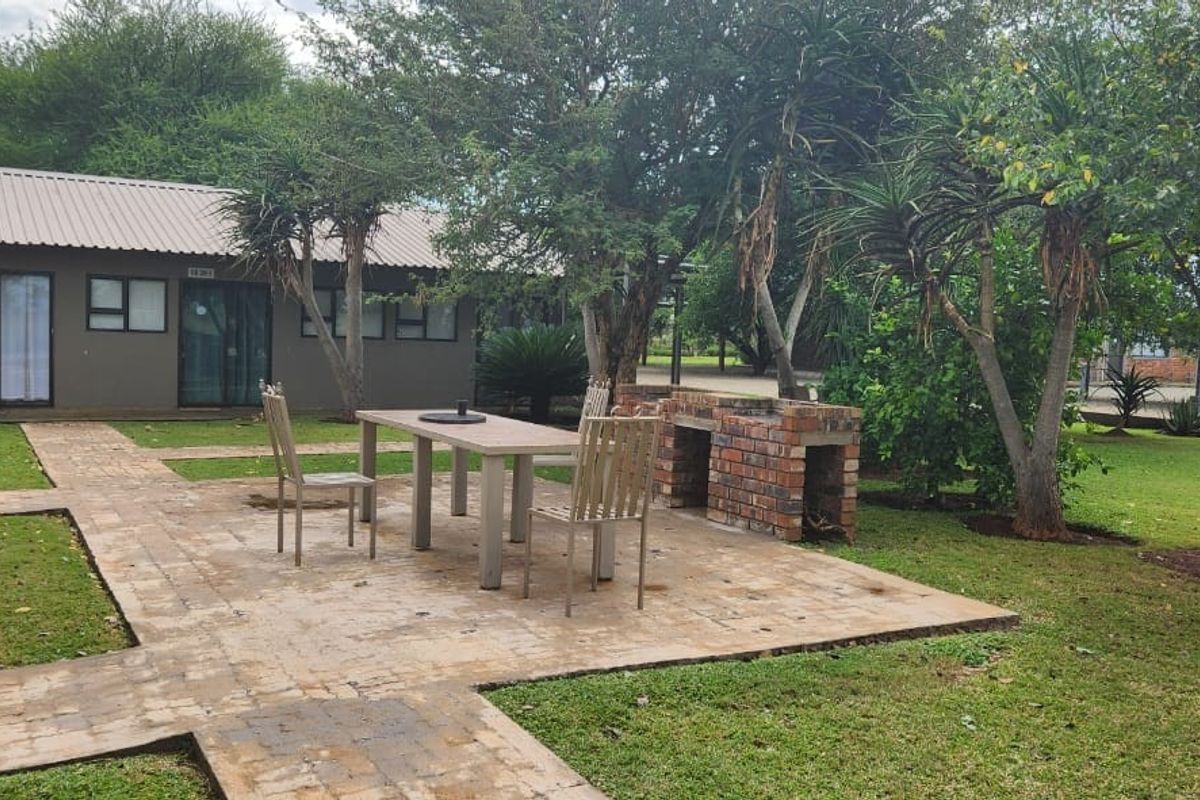 A well-maintained rear garden area featuring a paved patio with a built-in brick braai, surrounded by established trees and lawn.
