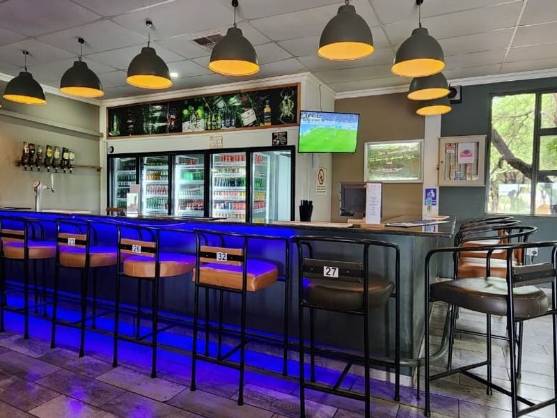 A well-equipped guest house bar area featuring a long built-in counter, commercial refrigeration, modern pendant lighting, and tiled flooring with direct outdoor access.