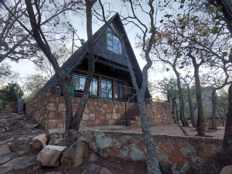 A unique A-frame house featuring stone cladding, a raised terrace, and large windows, set within a natural, wooded environment.