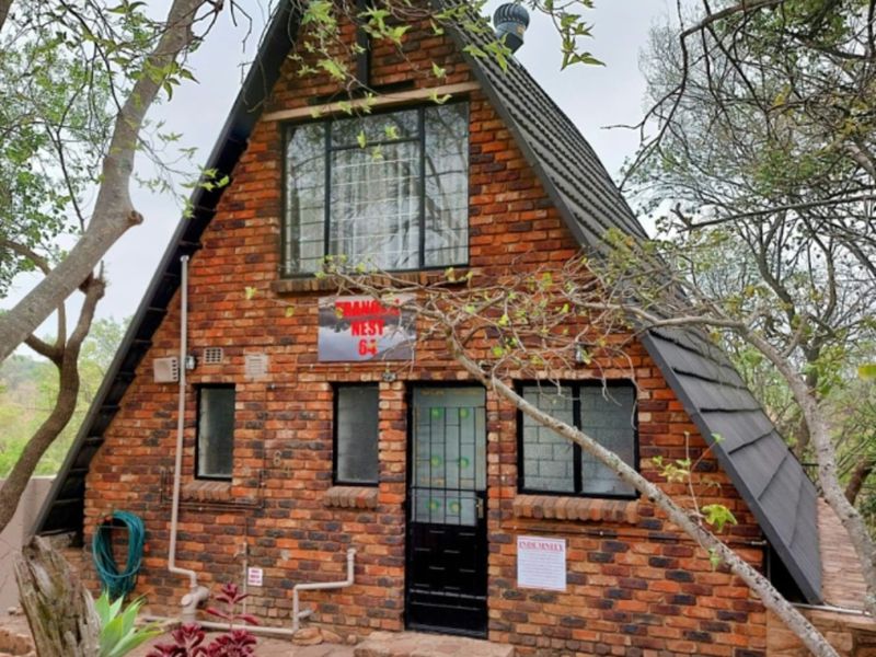 A charming A-frame house featuring a classic brick facade, tiled roof, and a stone-paved entrance area surrounded by mature trees.