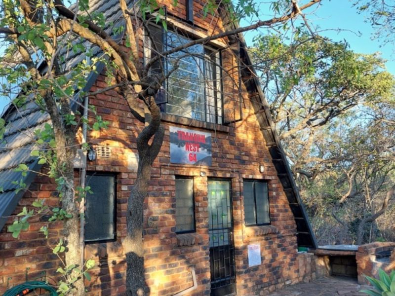 A charming A-frame face brick cottage featuring a stone-paved patio area and a built-in outdoor braai, nestled in a natural wooded setting.