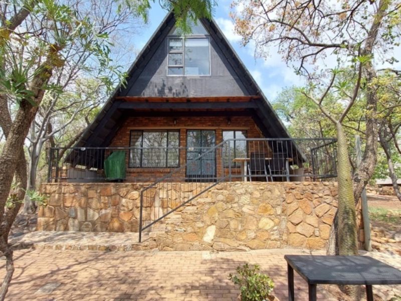 A charming A-frame house featuring stone cladding, a raised deck with metal railings, and a brick-paved entrance area surrounded by mature trees.