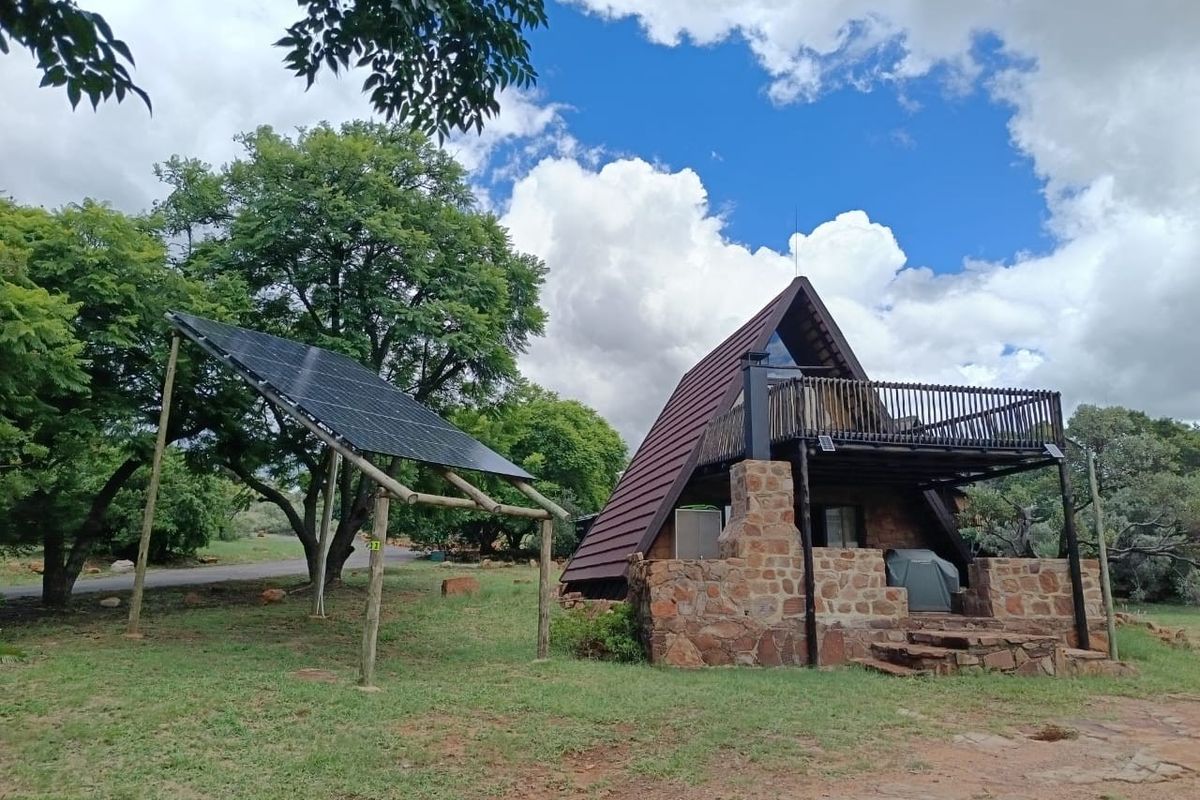 A rustic A-frame house featuring stone masonry, a timber balcony, and a ground-mounted solar panel array set in a natural garden landscape.